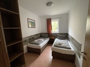 Een kleine, goed verlichte kamer met twee eenpersoonsbedden, een nachtkastje met een lamp, een raam met gordijnen en een wandplank.