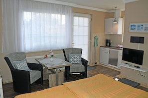 Una habitación de hotel moderna con una cama, dos sillones, una mesita con un florero, una kitchenette y un televisor.