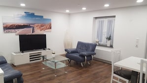 Ein modernes Wohnzimmer mit Flachbildfernseher, Glastisch, blauem Ledersofa, weißem Esstisch mit einem Stuhl und einem Fenster mit Vorhängen.