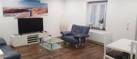 Una sala de estar moderna con un televisor de pantalla plana, una mesa de centro de vidrio, un sofá de cuero azul, una mesa de comedor blanca con una silla y una ventana con cortinas.