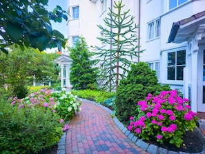 A garden pathway leading to a building with a white facade, surrounded by lush greenery and flowering plants.