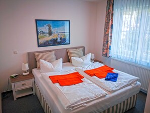 Un letto matrimoniale con lenzuola bianche, cuscini a righe e asciugamani arancioni e blu. Sopra il letto è appeso un quadro di un paesaggio.