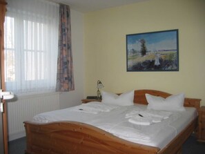 A bedroom with a wooden bed, white bedding, a painting on the wall, and a window with sheer curtains.