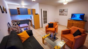 A living room with a sofa, two armchairs, a coffee table, a TV, and a kitchen area with cabinets and a refrigerator.