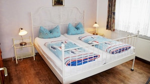 A double bed with a white metal frame, two matching pillows, and a blue blanket with a nautical design. There are two bedside tables with lamps, a small shelf, and a painting on the wall. - Ferienappartement 02 - Pension/holiday apartments in the Ostseebad Sellin (Sellin)