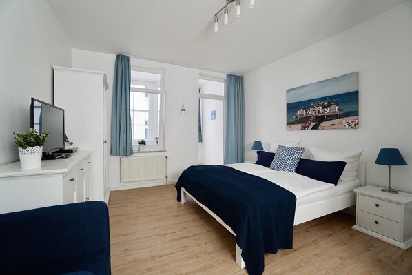 A bedroom with a bed, bedside tables, a TV, and a painting of a coastal scene. - 09 Holiday apartment Granitz (A) - Apartment house Haus Arkona (Sellin)
