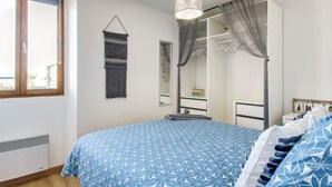 A bedroom with a large bed, a wardrobe, a hanging light fixture, and a window with curtains. - Basse Vallée by Interhome (Dinard)