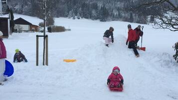 Une scène hivernale en plein air, enneigée, avec une luge, une personne portant une veste rouge et un enfant vêtu d’une combinaison de ski rose.