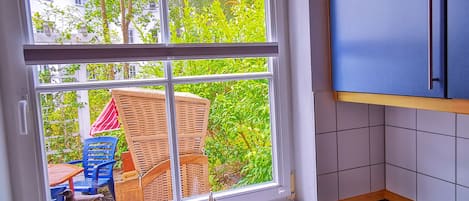 A kitchen with a large window, a dining area with outdoor furniture, and a view of greenery and buildings outside.
