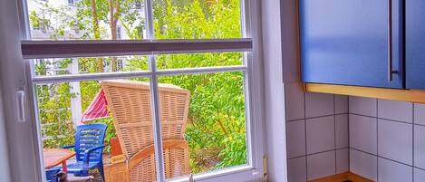A kitchen with a large window, a dining area with outdoor furniture, and a view of greenery and buildings outside.