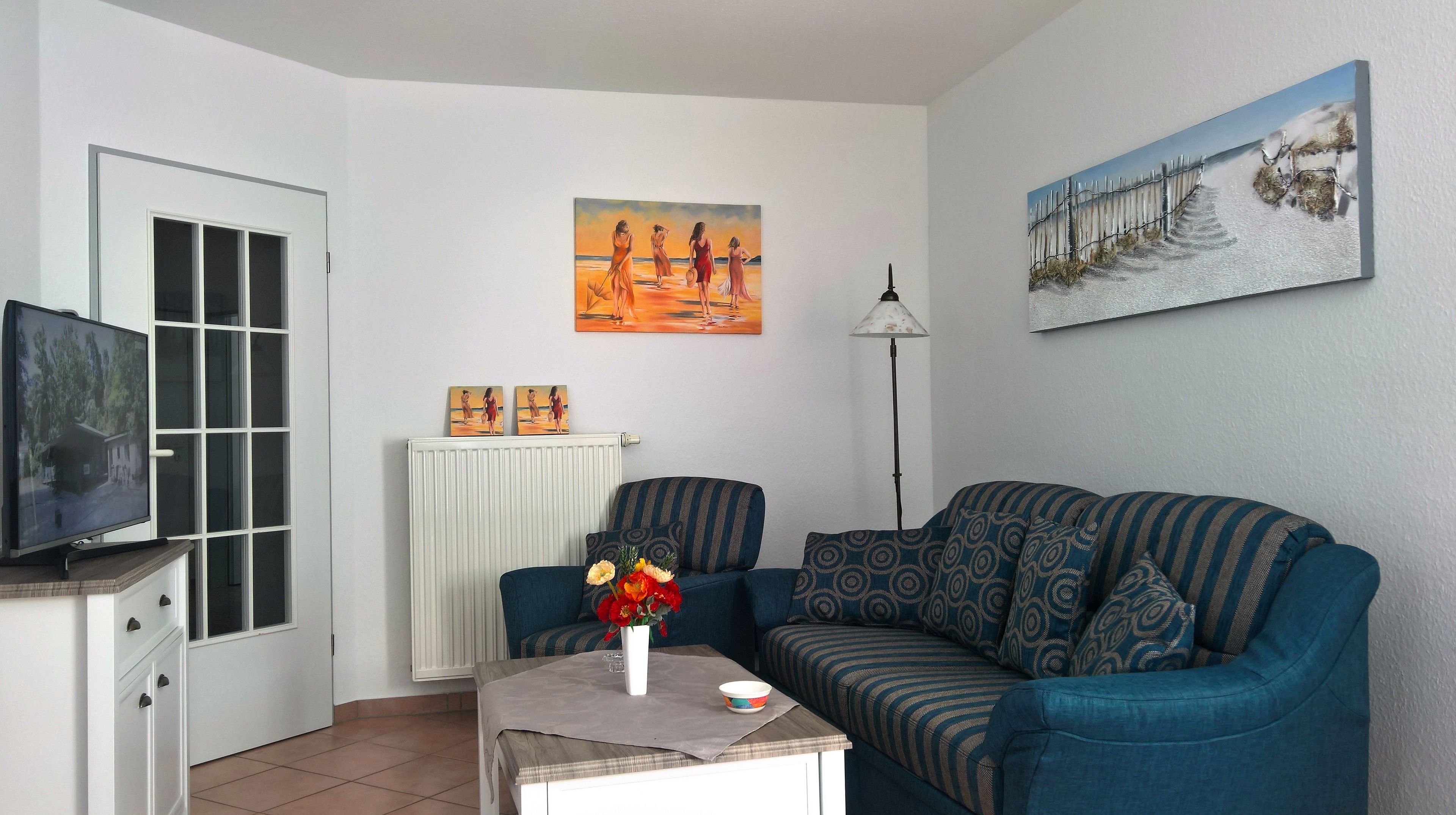 Een woonkamer met een televisie, een bank, een salontafel met een bloevas en twee schilderijen aan de muur.