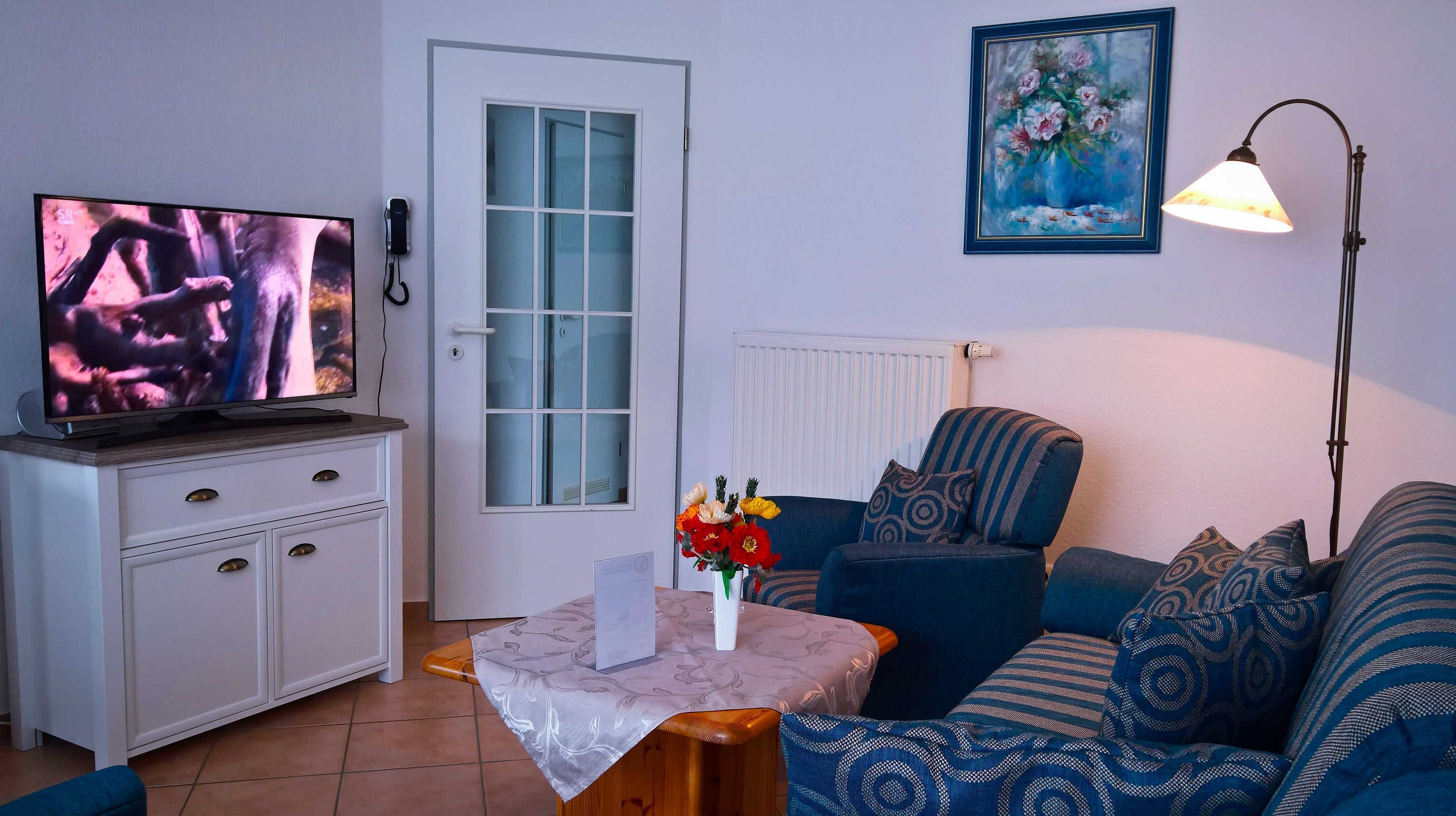 A living room with a television, a white cabinet, a blue sofa, a small table with a vase of flowers, and a painting on the wall.
