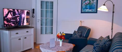 Uma sala de estar com televisão, um armário branco, um sofá azul, uma mesa pequena com um vaso de flores e um quadro na parede.