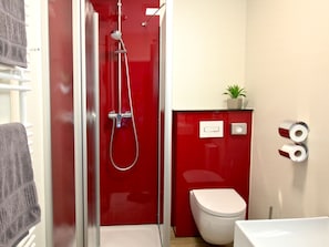 A compact bathroom with a red wall, a white toilet, and a shower. There is a towel rack and a small potted plant on the wall.