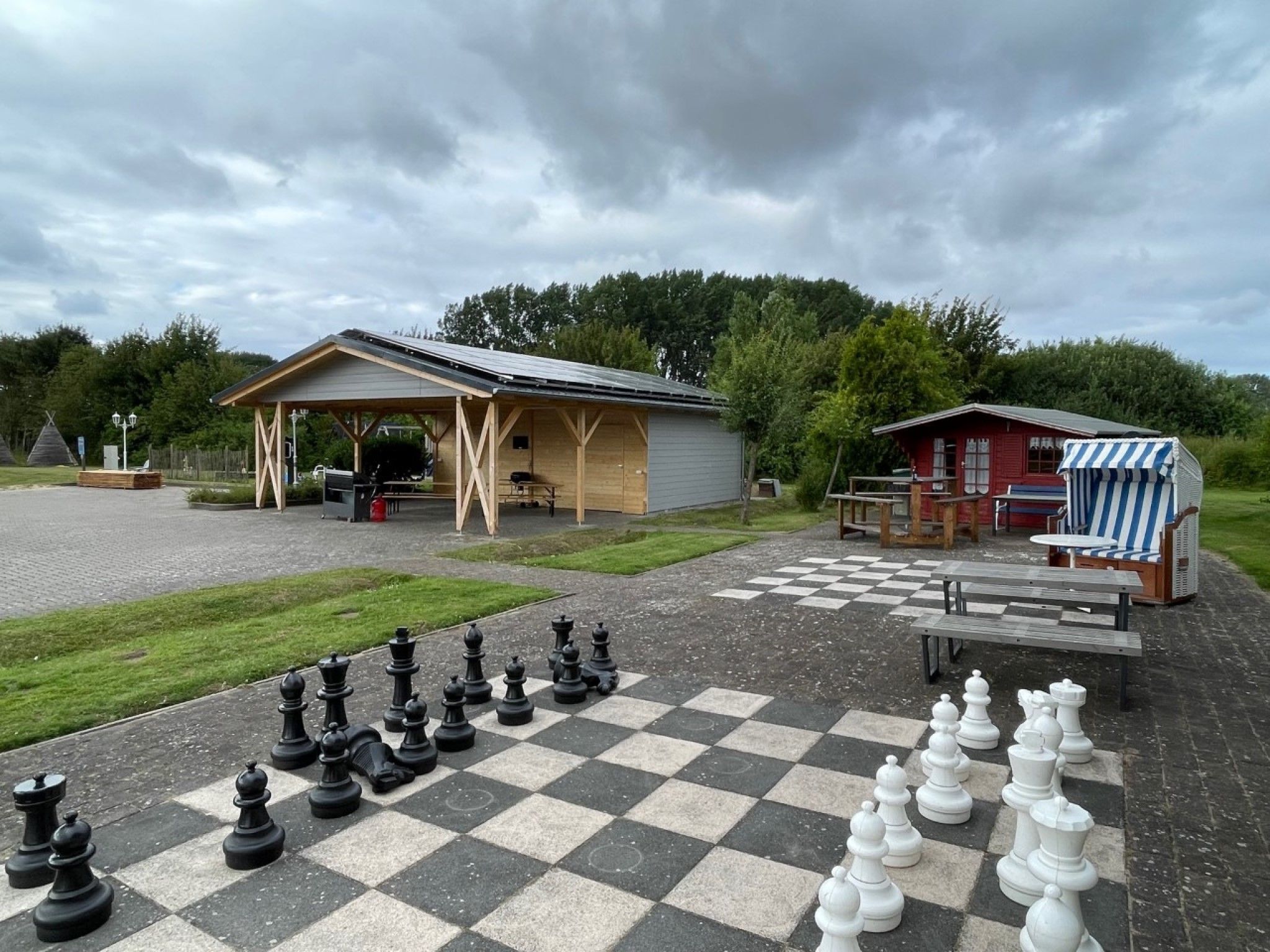 Ett stort utomhus-schackbräde placerat på en belagd yta, med en överbyggd paviljong och en liten röd byggnad i bakgrunden.