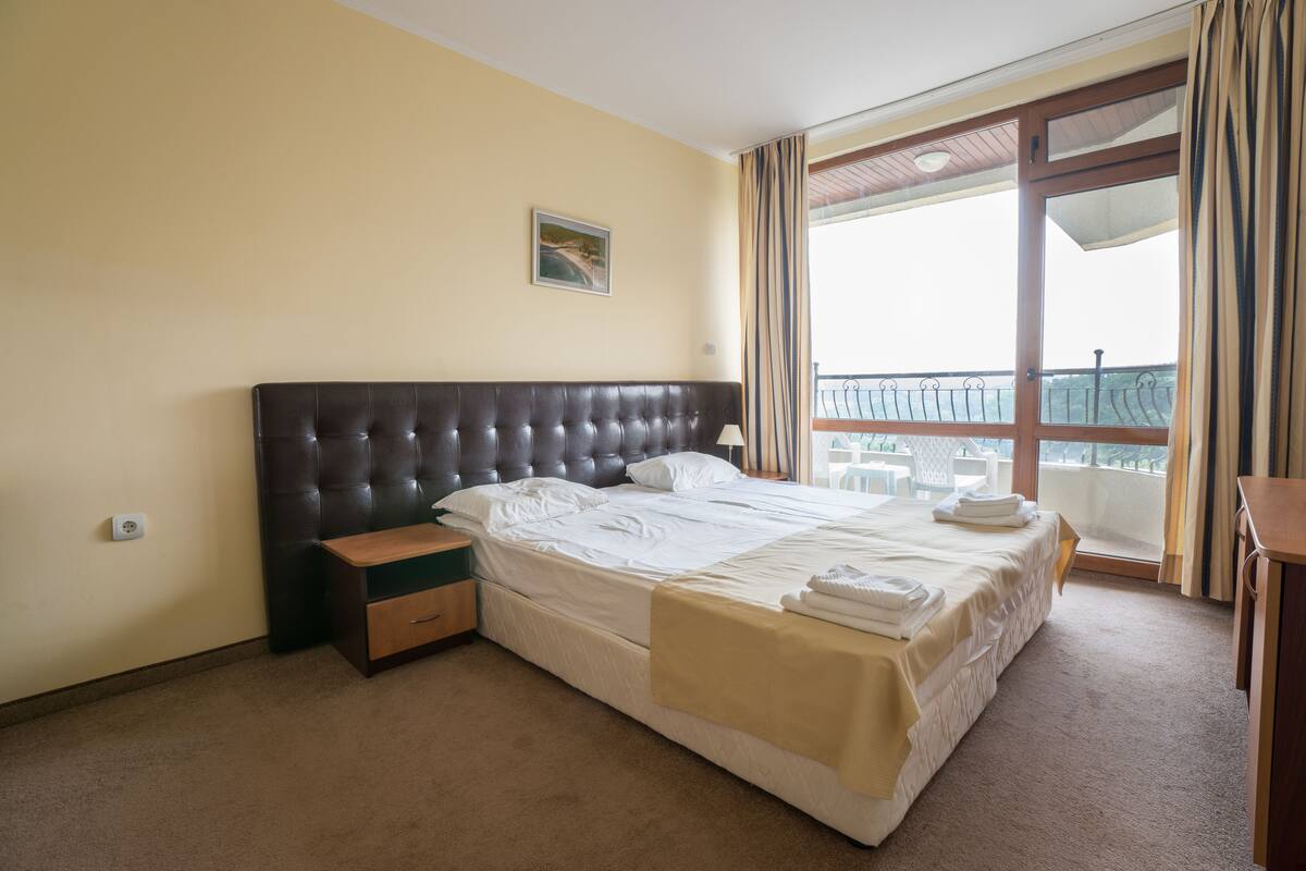 A bedroom with a large bed, a nightstand, a window with curtains, and a balcony view.