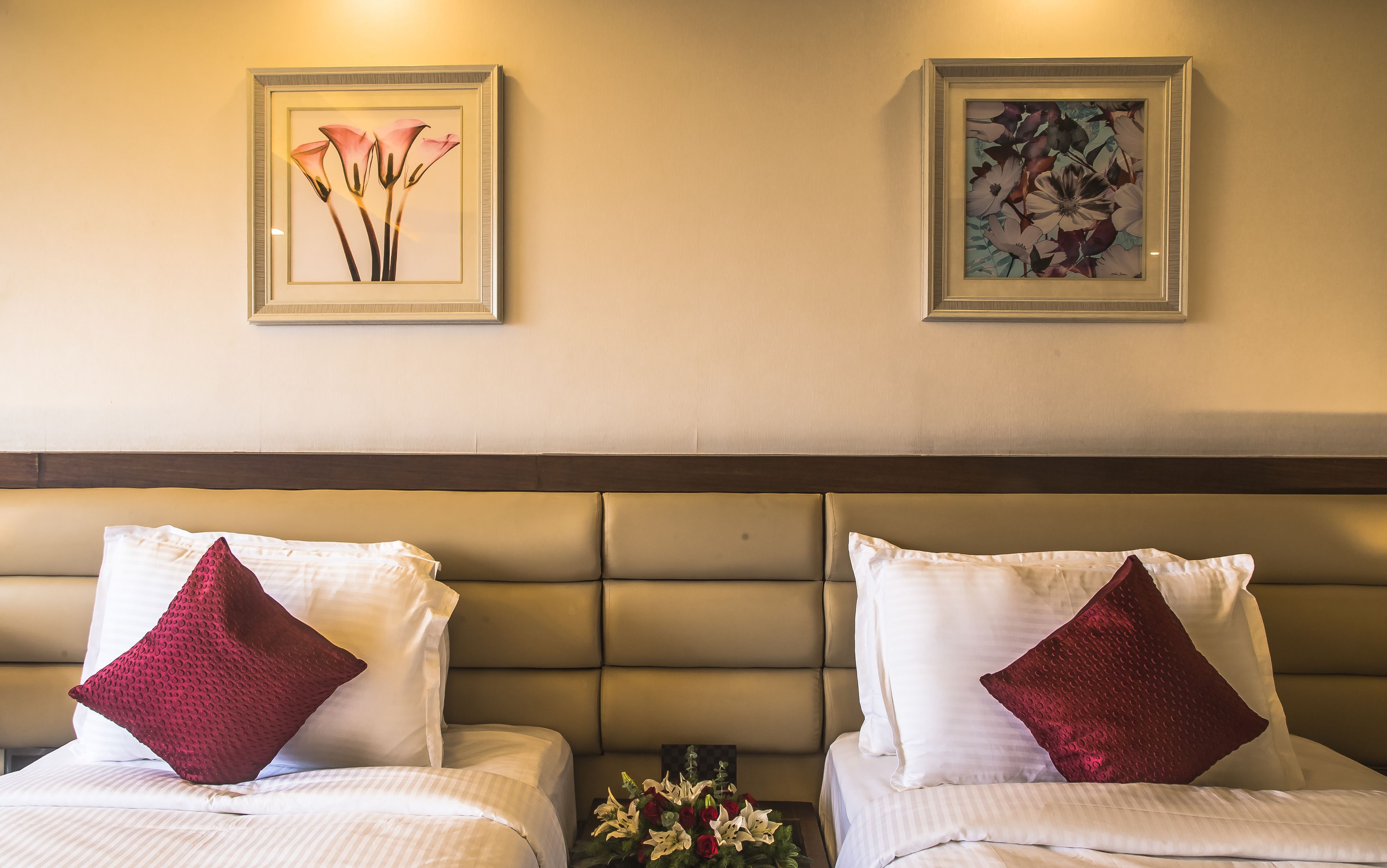 A hotel room with two beds, each with white bedding and a red pillow. There are two framed floral paintings on the wall above the beds.