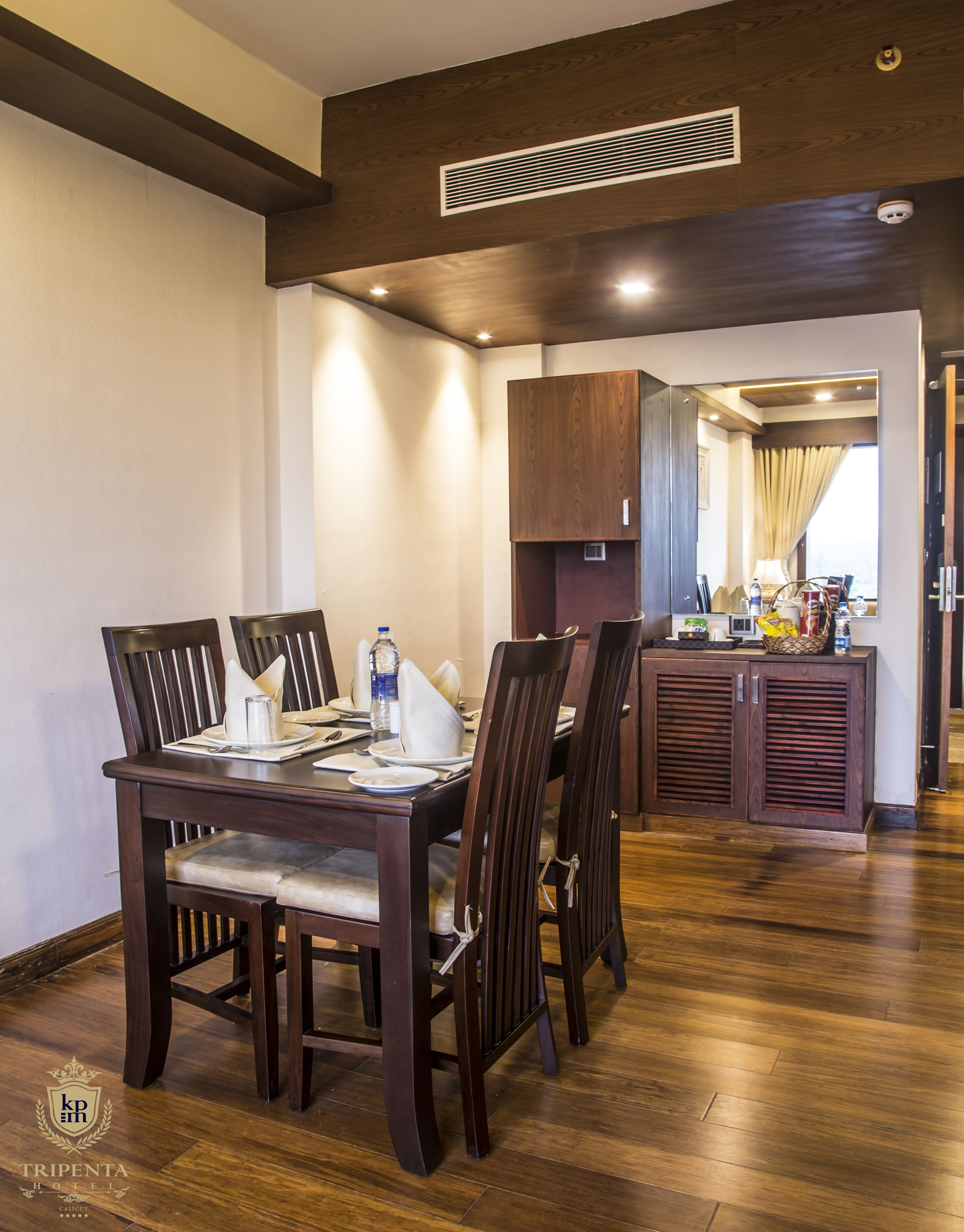 Un comedor con una mesa de madera dispuesta para cuatro, flanqueada por sillas de madera oscura. Se ve un área de cocina al fondo, con armarios de madera y una ventana con cortinas.
