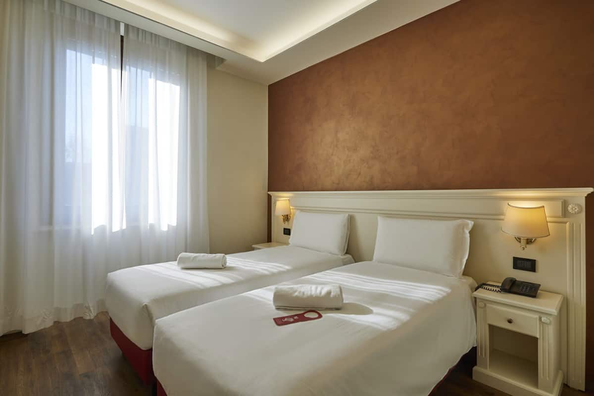 A hotel room with two beds, a nightstand with a lamp, and a view of the outdoors through sheer curtains.