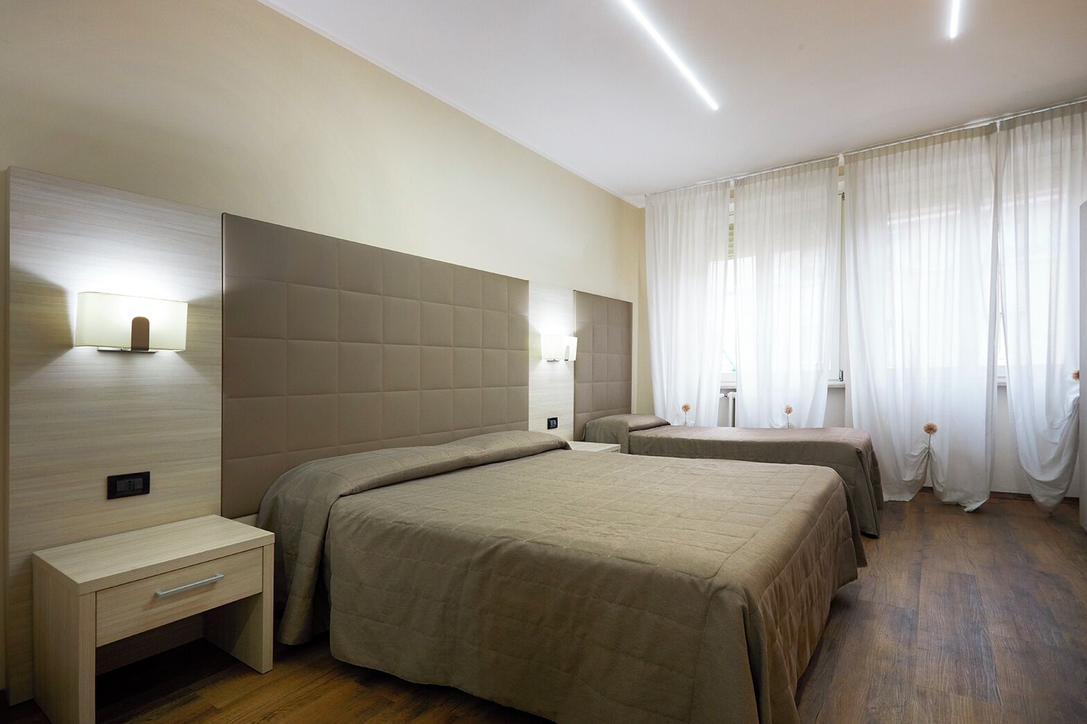 A hotel room with a large bed, bedside tables, a nightstand, a wall-mounted lamp, and a window with sheer curtains.