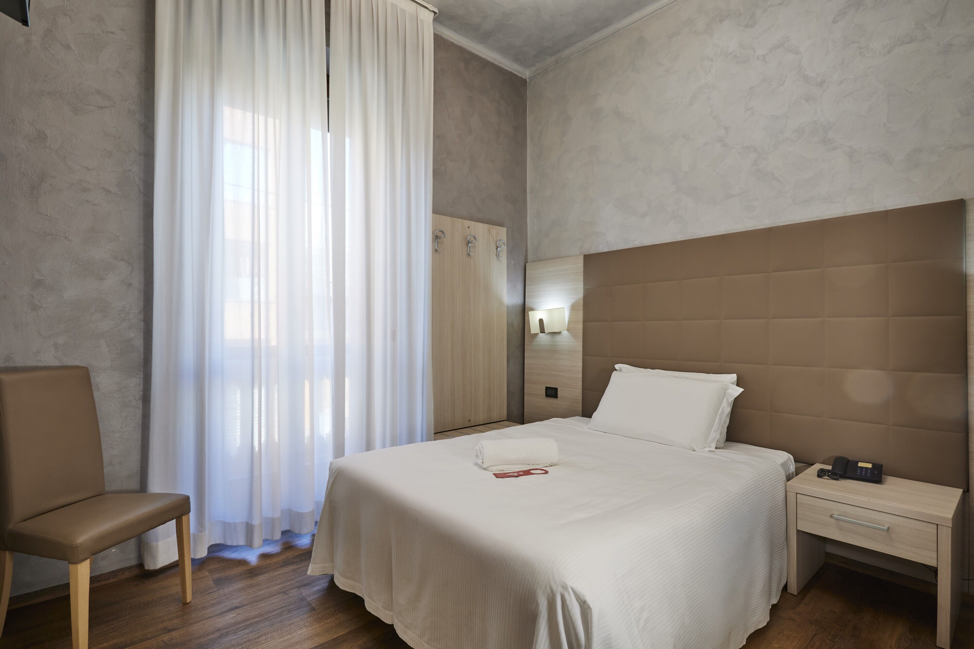 A hotel room with a large bed, a bedside table with a phone, a chair, a wardrobe, and a window with sheer curtains.