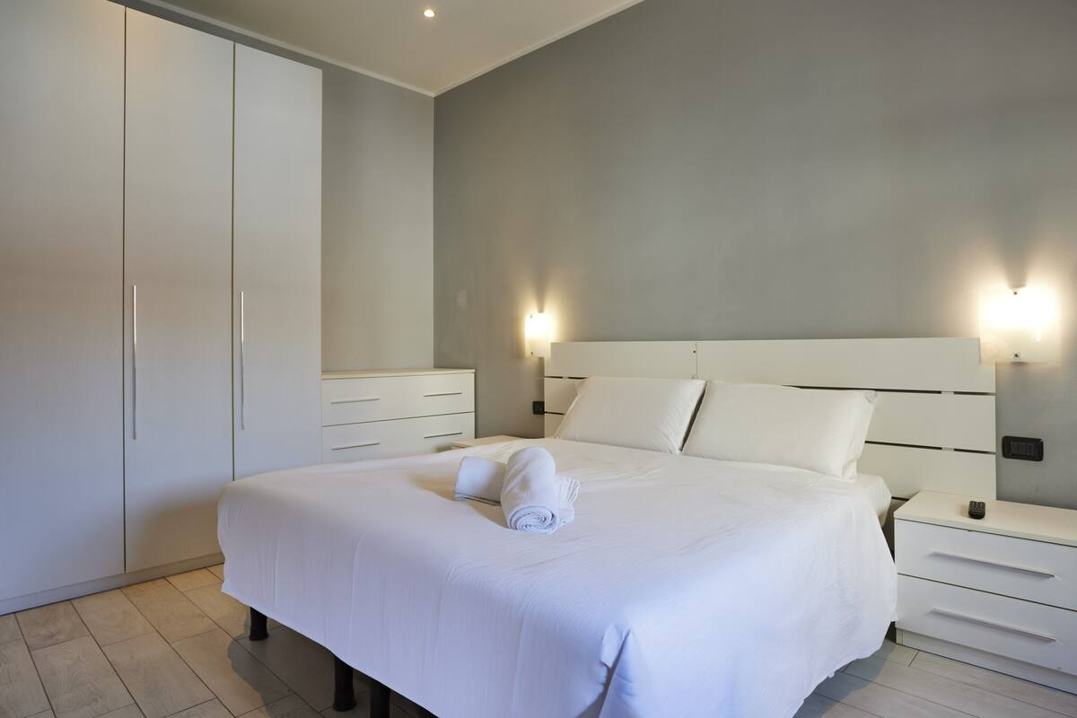 A bedroom with a large bed, white bedding, a nightstand with a lamp, and a wardrobe.