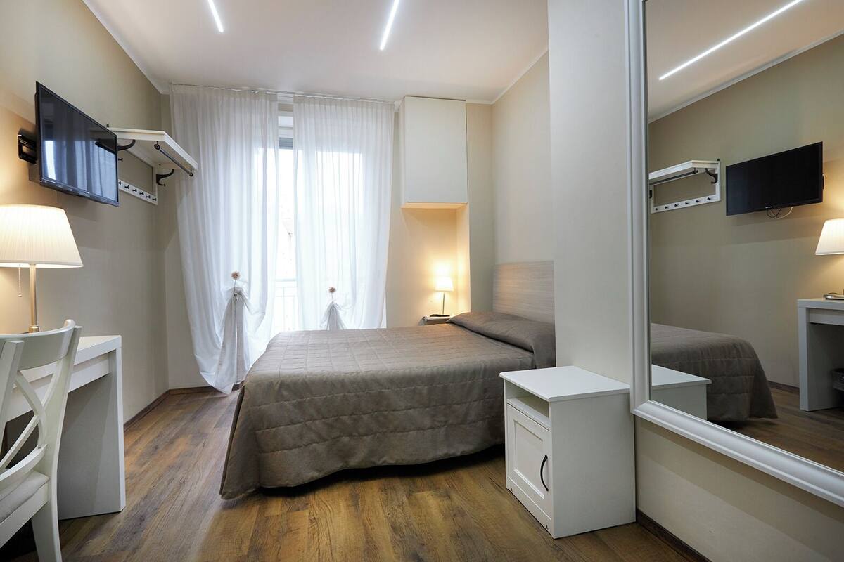 A bedroom with a bed, bedside tables, a desk, a chair, a mirror, and a television.