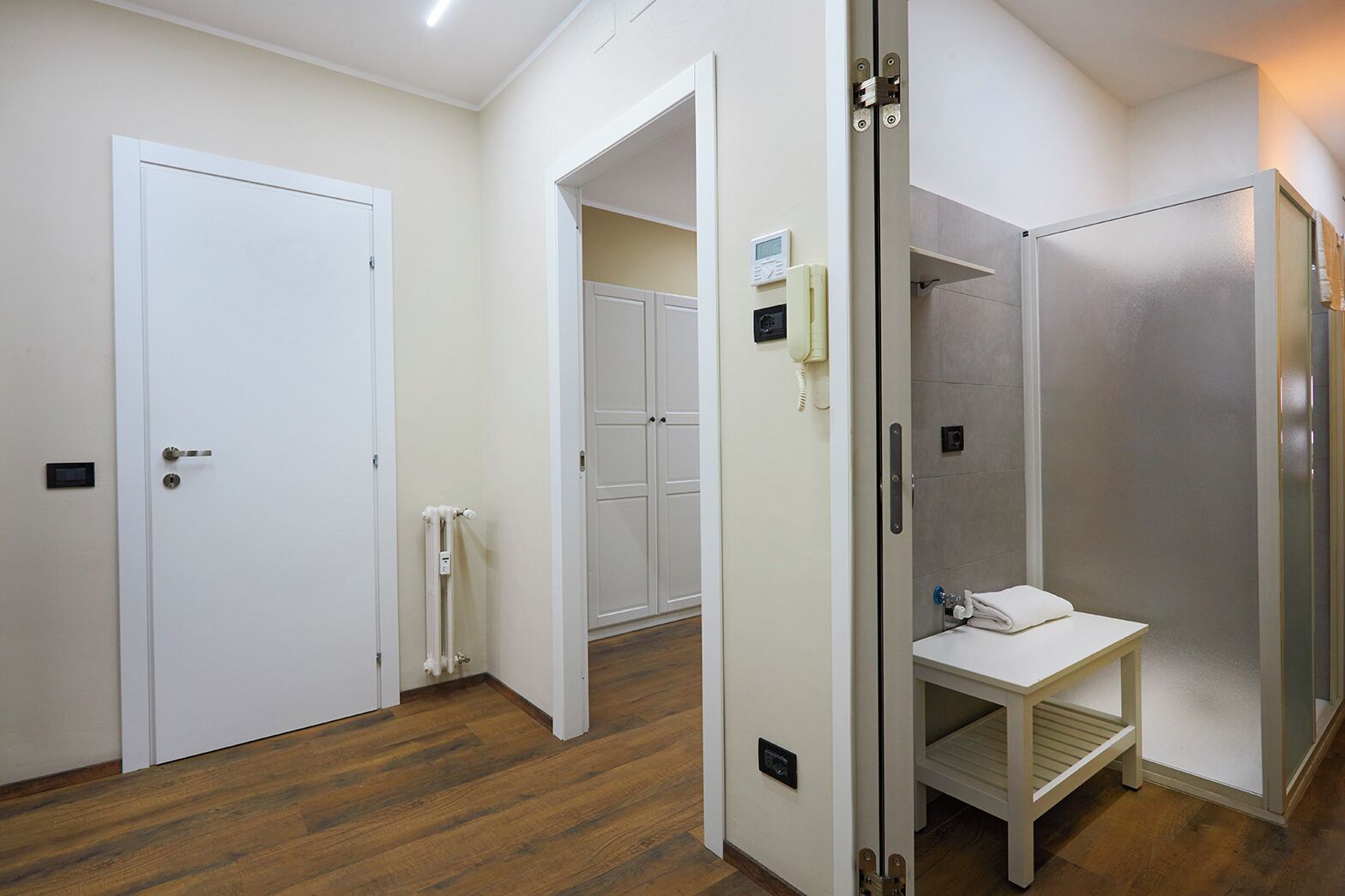 Ein Hotelzimmer mit Holzboden, einer weißen Tür, einer weißen Wand, einem weißen Schrank und einem weißen Handtuch auf einem kleinen Tisch.