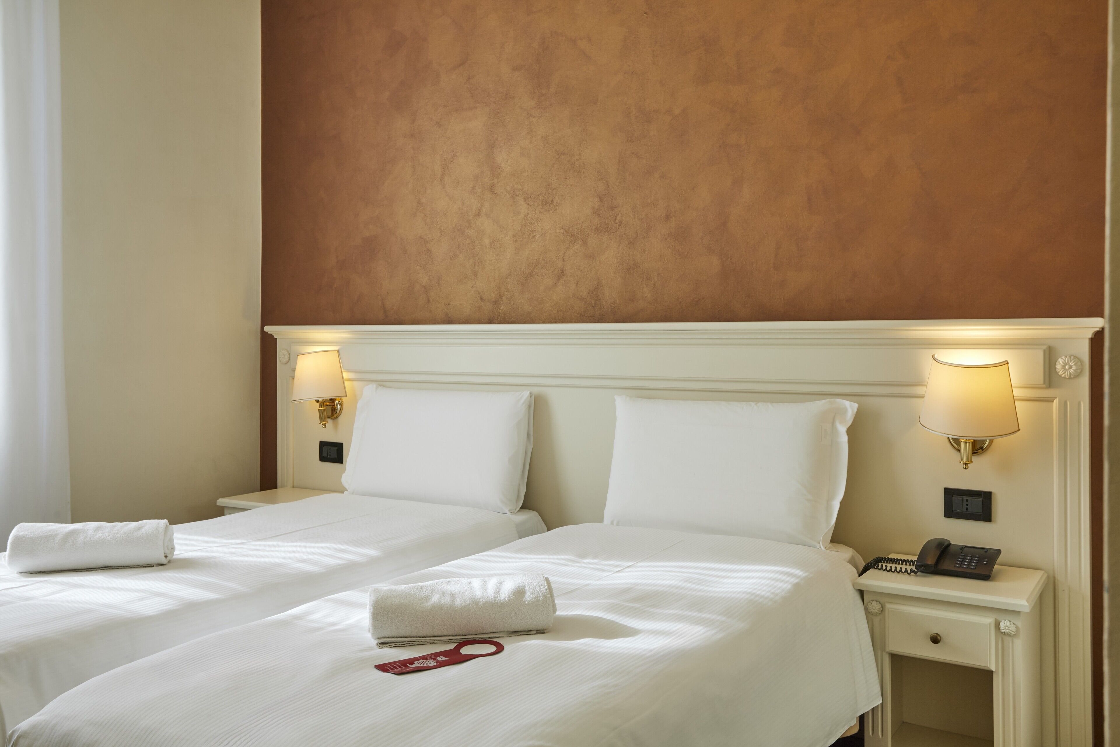 A hotel room with two beds, white linens, a red towel, a phone on the nightstand, and wall-mounted lamps.