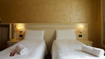 A hotel room with two beds, each with a white bedspread and pillows, a nightstand with a lamp, and a red object on the bed.