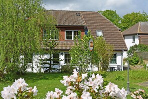 Een tweelaags huis met een pannendak, omgeven door groen en bloeiende planten.