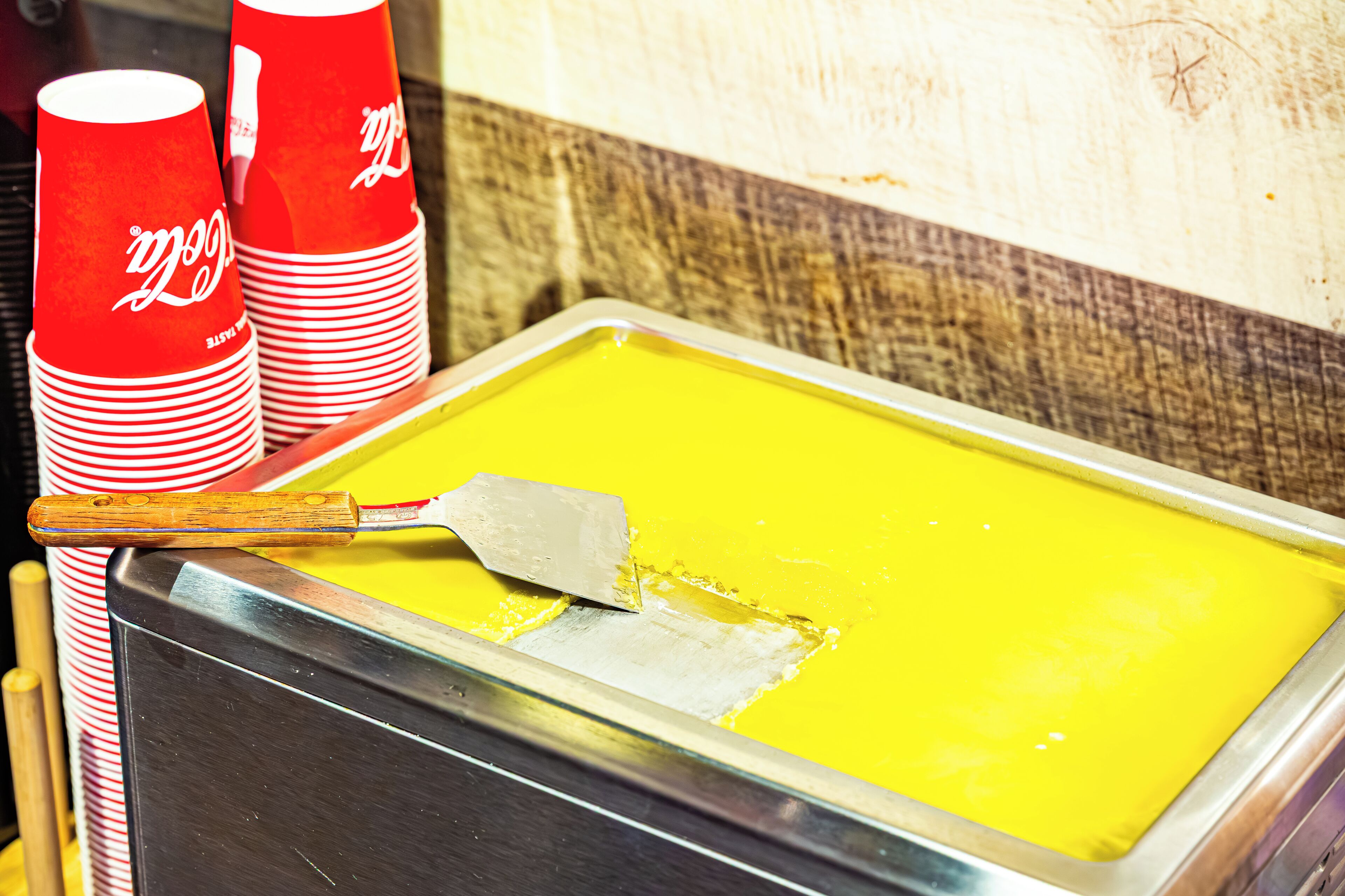 A yellow substance in a tray with a spatula, red and white Coca-Cola cups stacked beside it.