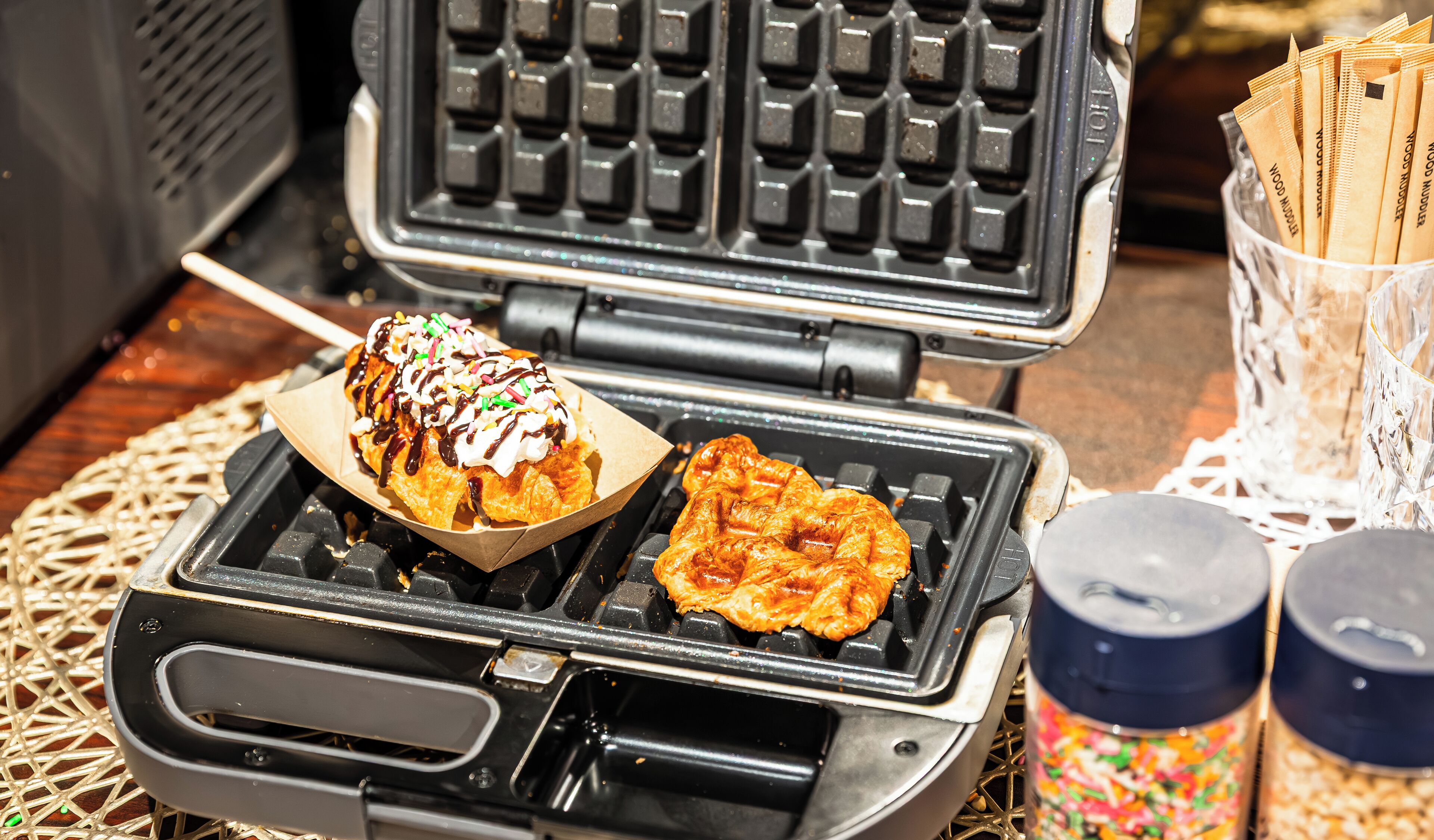 A waffle iron with two waffles, one topped with whipped cream and sprinkles, and a container of waffle mix.