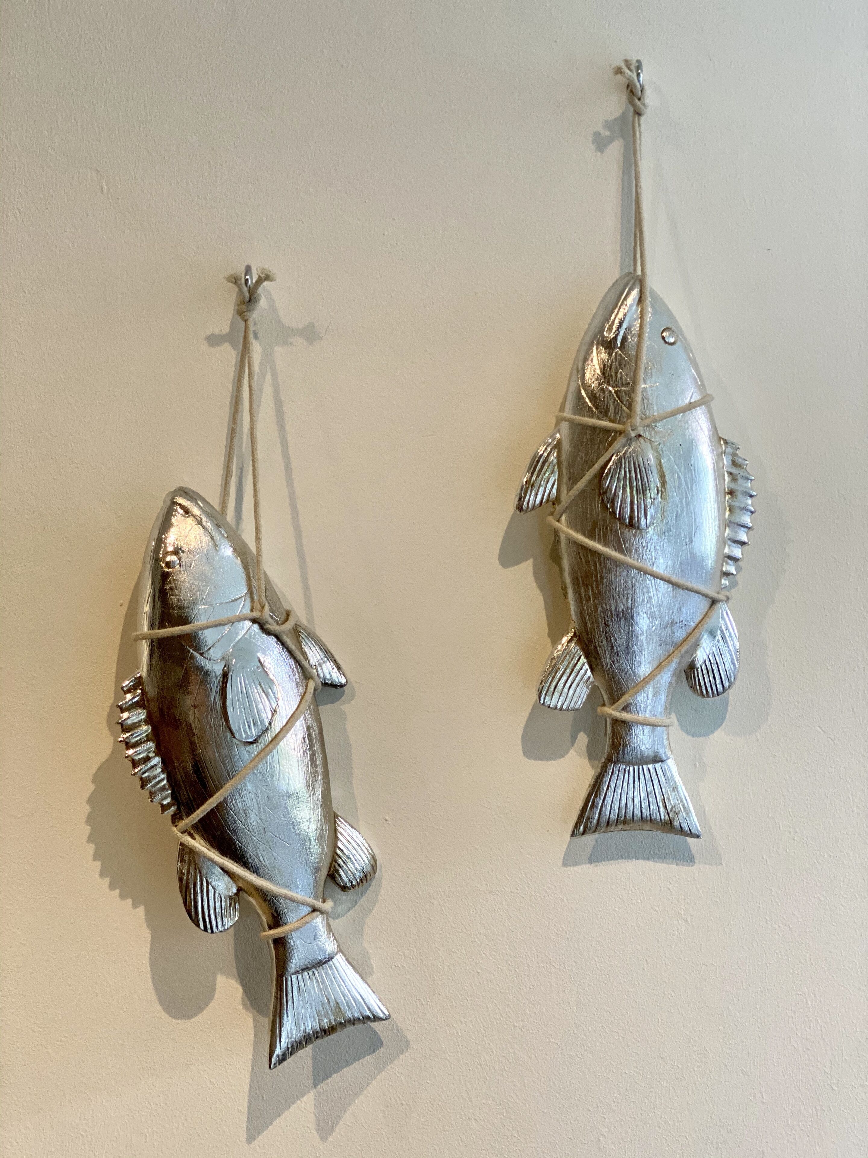 Two metallic fish-shaped wall decorations with strings, mounted on a plain wall.