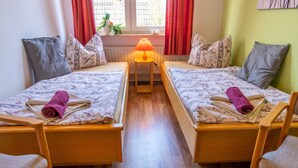 Et værelse med to enkeltsenge, et lille bord med lampe og et vindue med røde gardiner.