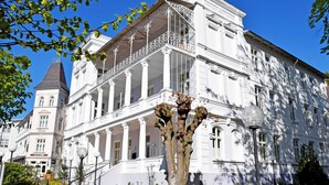 A large white building with a prominent balcony and columns, surrounded by greenery and street lamps.