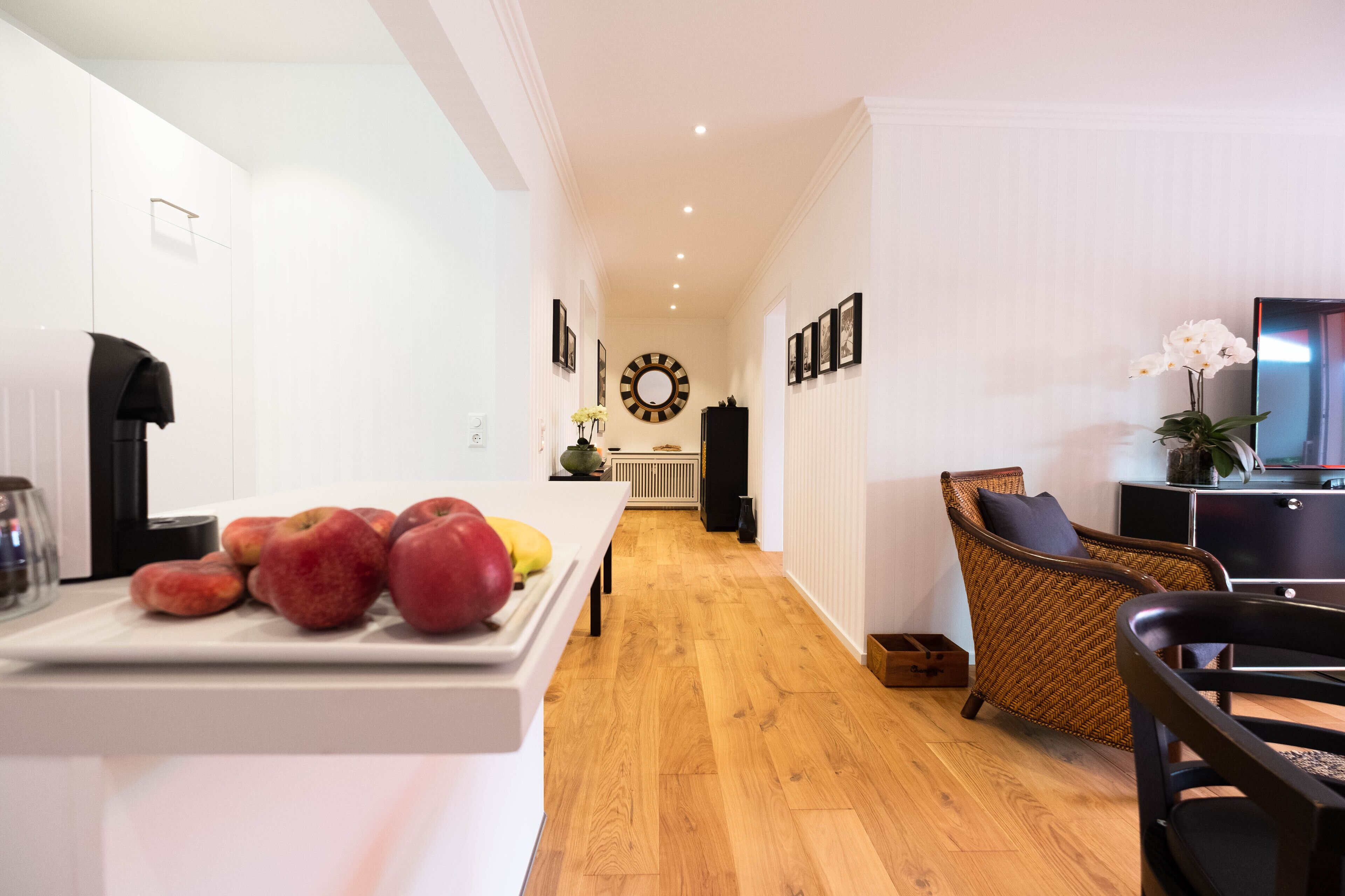 A modern kitchen with a wooden floor, white cabinetry, and a dining area with wicker furniture.