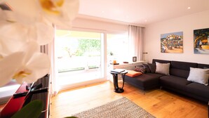 A modern living room with a dark sofa, a small side table, and a large window offering a view of greenery. - Beautiful apartment in a dream location near the Festival Hall (Baden Baden)