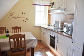 Cuina amb armaris blancs, microones i cafetera. Hi ha una finestra amb cortina i una taula amb cadires.