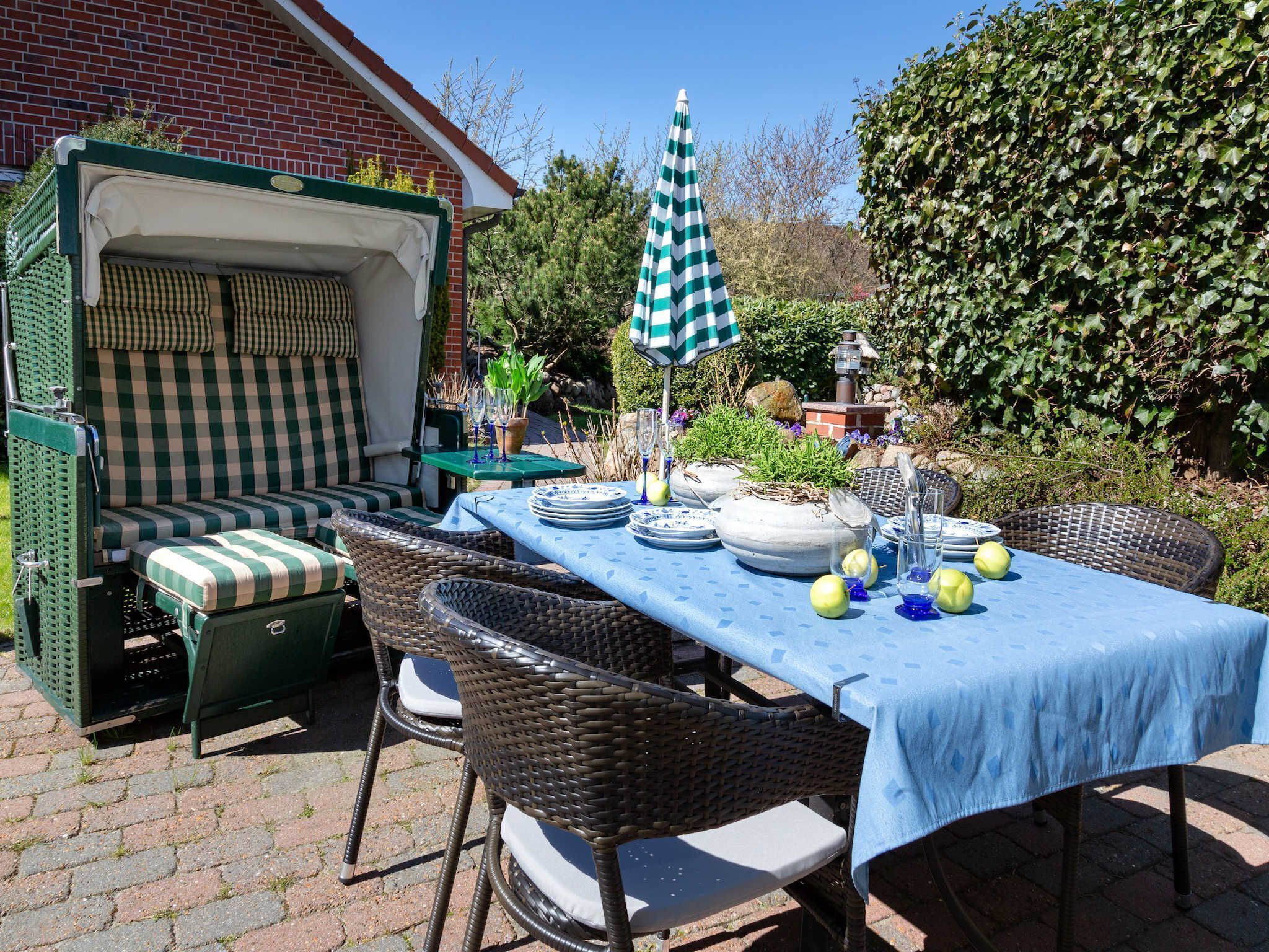 Un patio exterior con un sofá a cuadros, sillas de mimbre, una mesa dispuesta con platos, vasos y un paraguas verde.