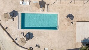 An aerial view of a swimming pool with surrounding lounge chairs and umbrellas, a covered seating area, and a building with a flat roof.