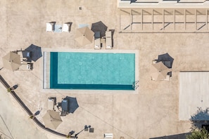 Vista aérea de una piscina con sillas de descanso y sombrillas alrededor, un área de estar cubierta y un edificio con techo plano.