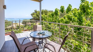 A balcony with a round table and chairs, overlooking a scenic view of the sea and lush greenery.