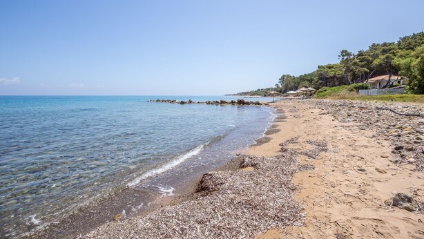 A beach with clear water, pebbles, and a few houses in the background.