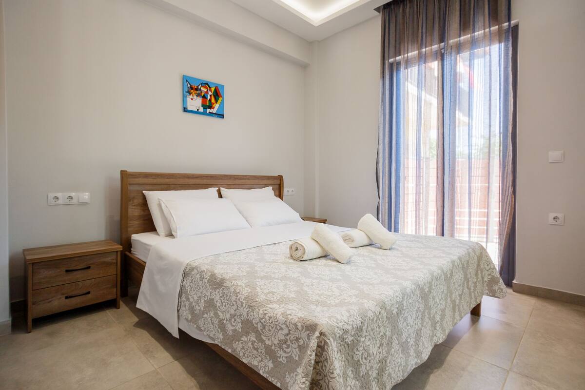Una cama bien hecha con un edredón y almohadas estampadas, una mesita de noche de madera con cajones, una pequeña obra de arte en la pared y una ventana con cortinas.