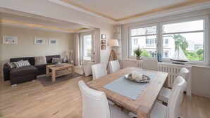 Een moderne woonkamer met een bank, salontafel, eettafel en stoelen. Er hangen ingelijste foto's aan de muur, een lamp en er is uitzicht op gebouwen door het raam.