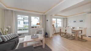 Een ruime woonkamer met een bank, salontafel, eettafel en stoelen. Grote ramen met gordijnen bieden uitzicht op buiten, en het plafond heeft zichtbare balken.