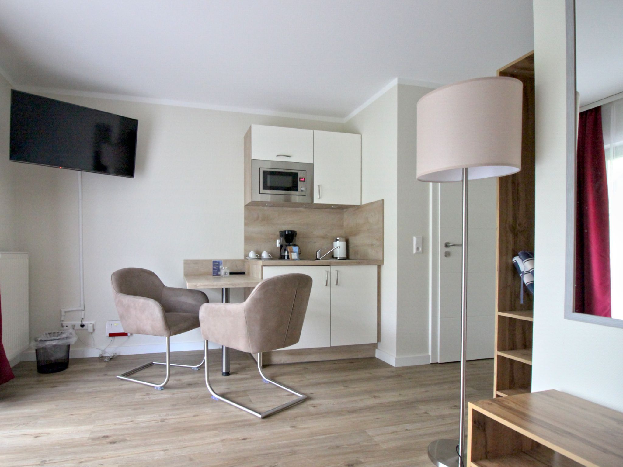 Ein modernes Zimmer mit Flachbildfernseher, einer Küchenzeile, zwei Stühlen, einer Stehlampe und einem Holzregal.