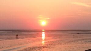 Uma praia ao pôr do sol, com mar calmo e algumas pessoas caminhando.