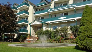 Um hotel com uma placa de destaque que diz "Strandhotel Steiner". Apresenta um pátio central com uma fonte, rodeado por vegetação bem cuidada e um conjunto de sebe aparado.